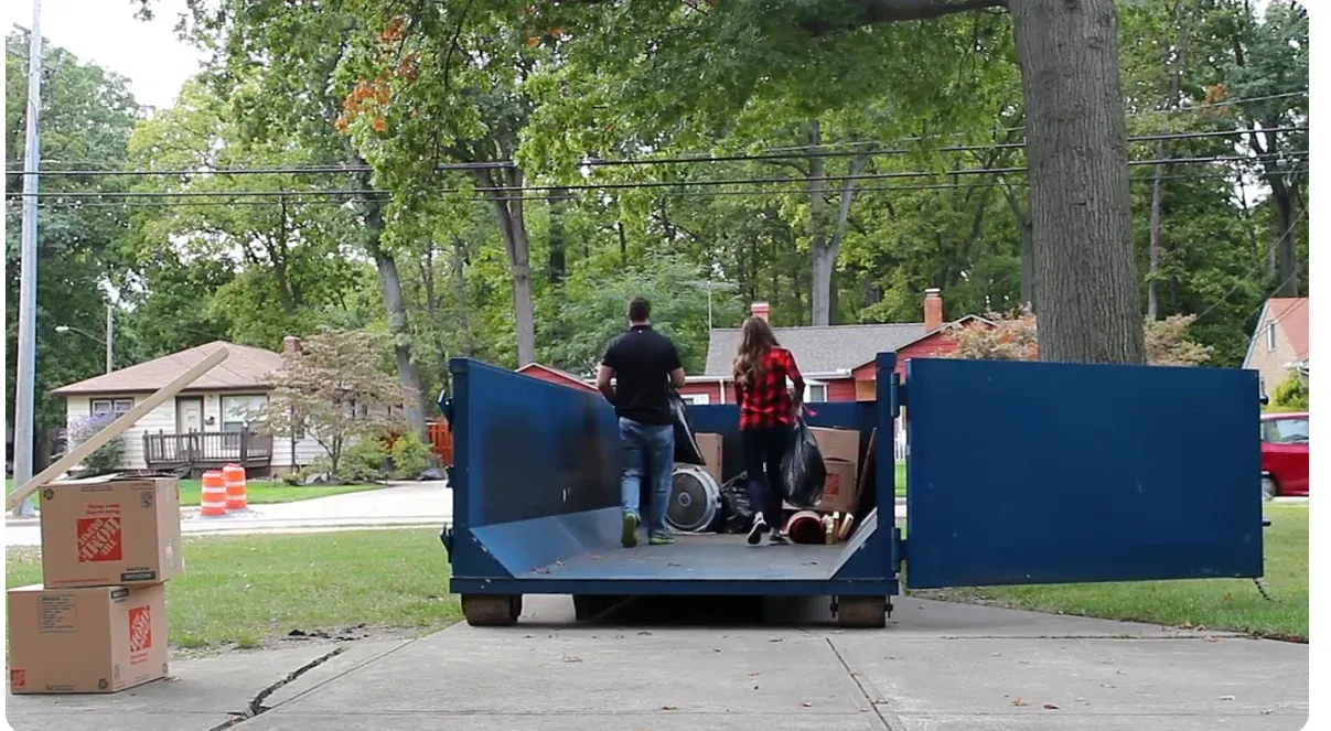 Two people carrying household items into a roll-off dumpster through its open swing door on a driveway